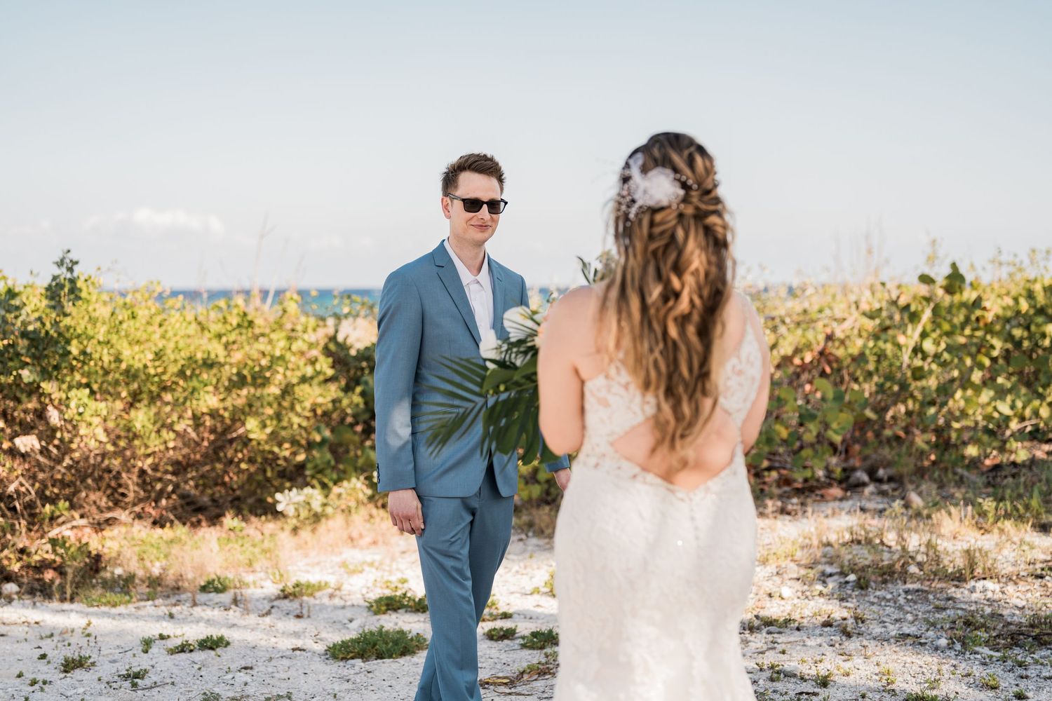 Groom in blue suit smiles at bride during romantic first look on sun-drenched beach.