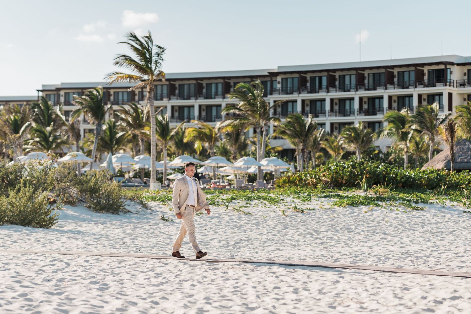 Groom in tan suit walks along white sandy beach with luxury resort hotel in background.