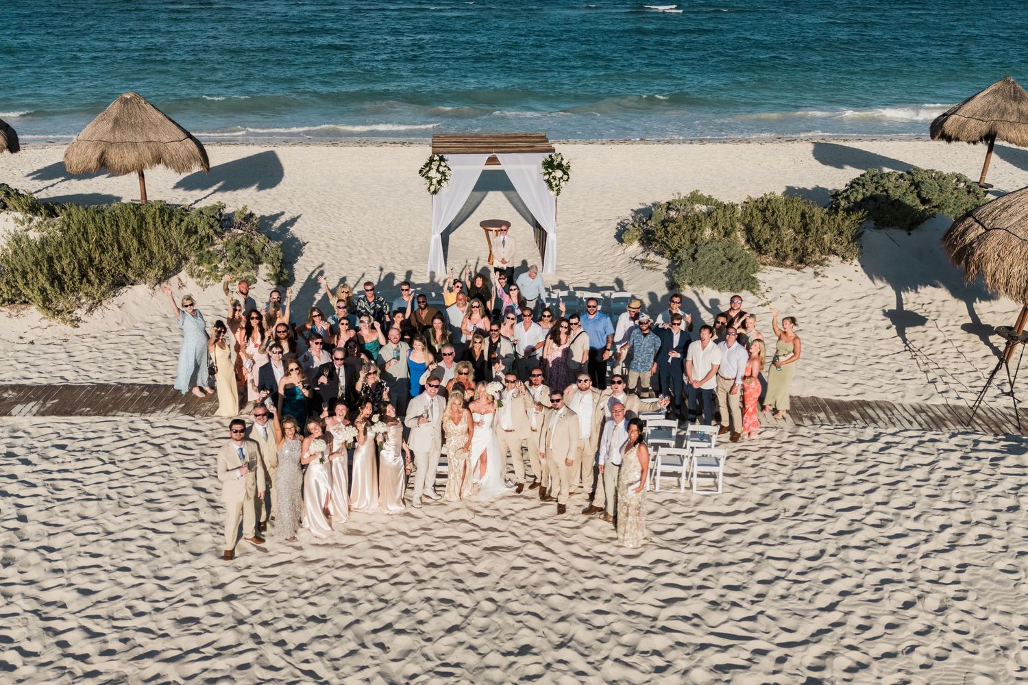 Large wedding group photo on sandy beach with ceremony arch and ocean in the background.