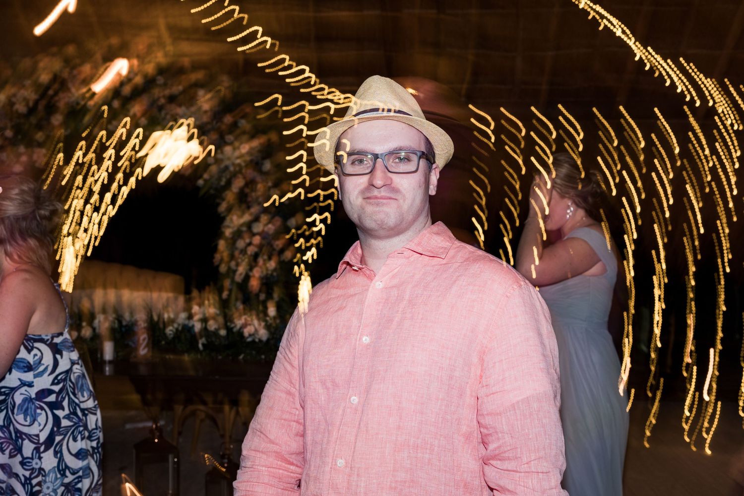 Wedding guest in pink shirt and straw hat poses under spinning fairy lights at reception.