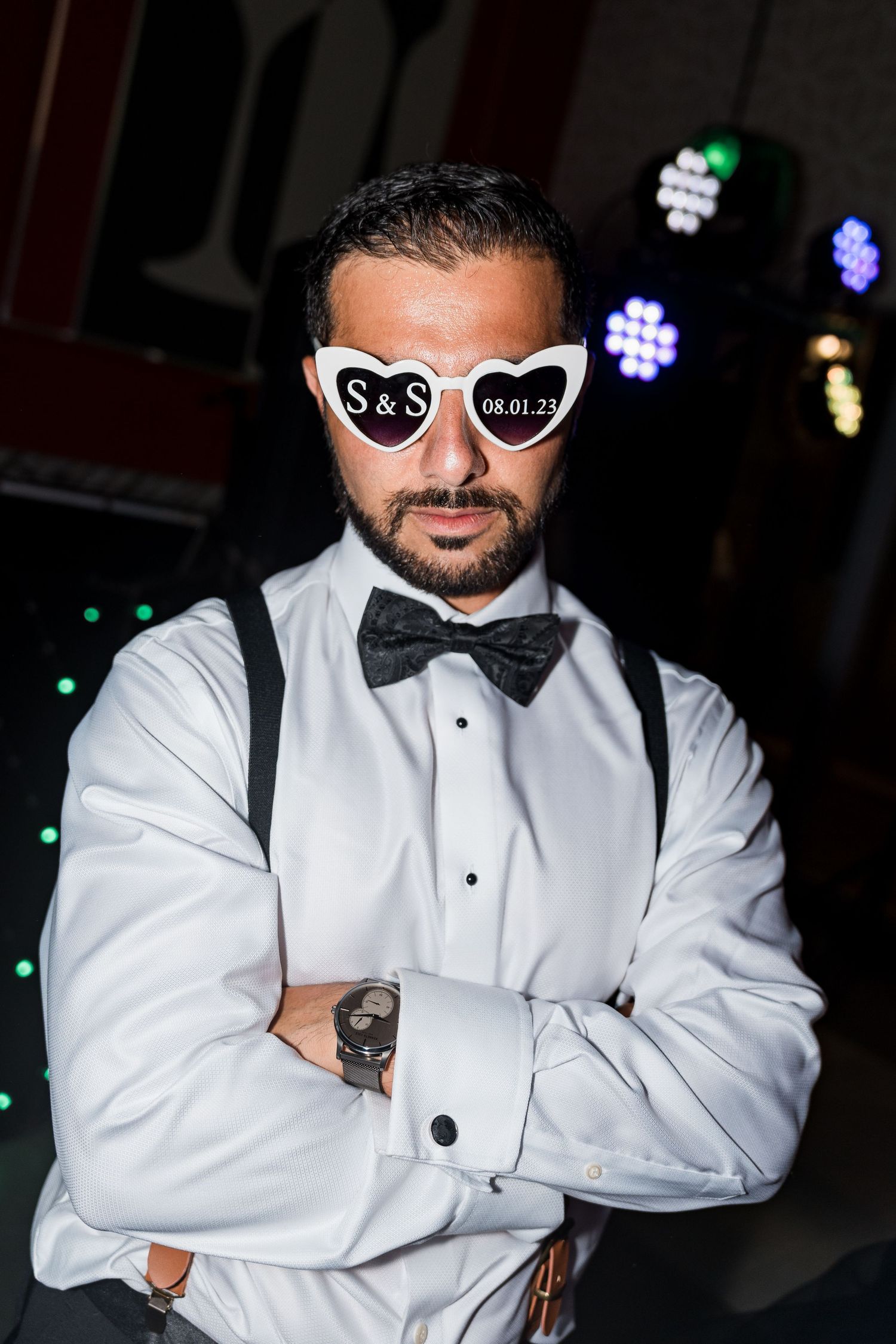 Man in tuxedo and bow tie wearing S&S heart-shaped wedding sunglasses poses with arms crossed.