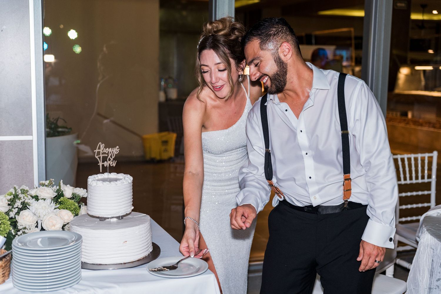 Smiling bride and groom cutting their two-tier white wedding cake together.