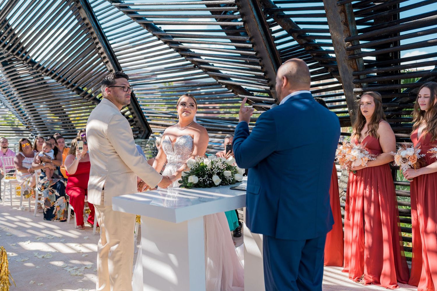 Bride and groom exchange vows at outdoor altar with officiant and bridesmaids in red.