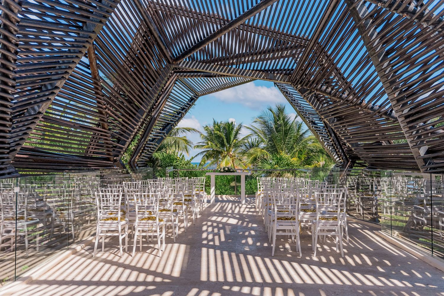 Open-air bamboo wedding ceremony venue with clear acrylic chairs and tropical palm tree views.