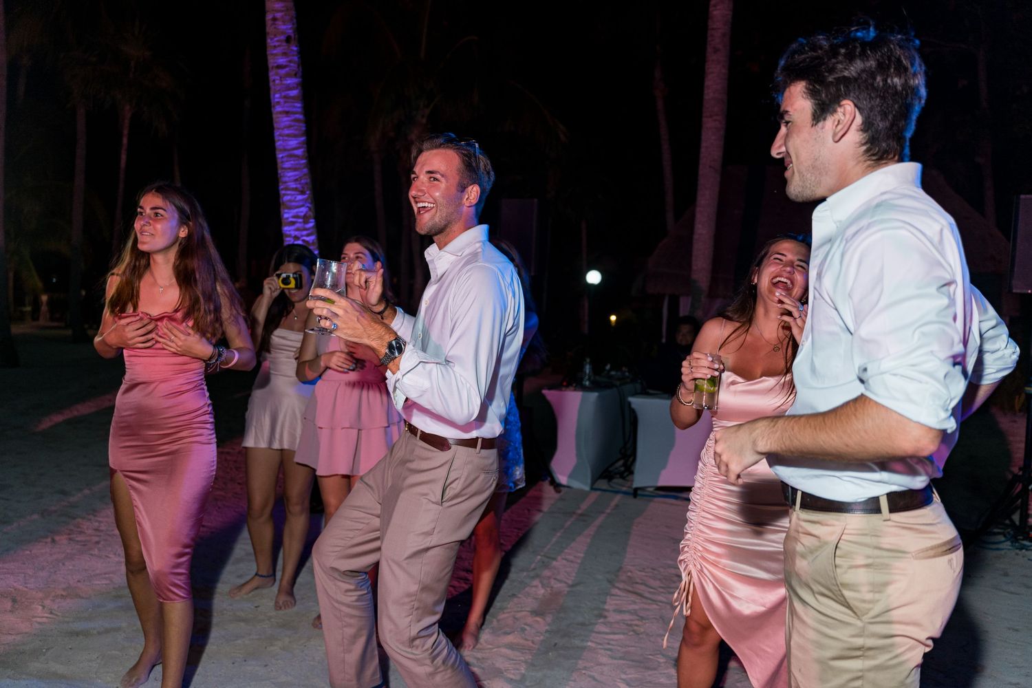 Elegantly dressed wedding guests in pink dresses laugh and dance at a beach reception at night.