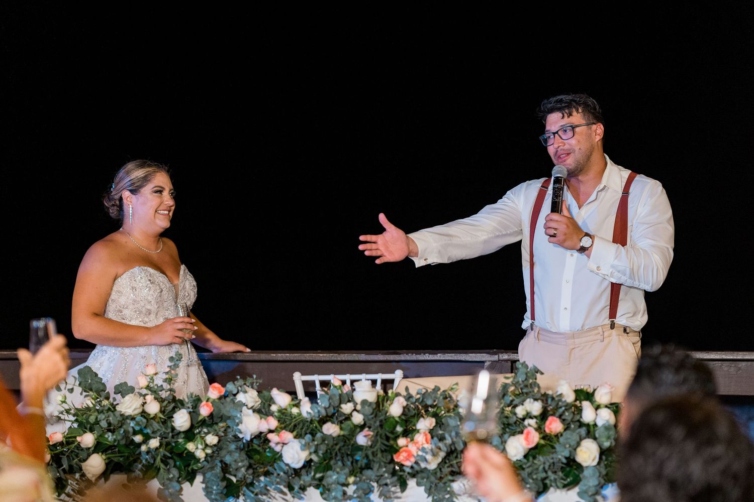 Groom gesturing toward smiling bride during wedding reception speech at decorated outdoor venue.