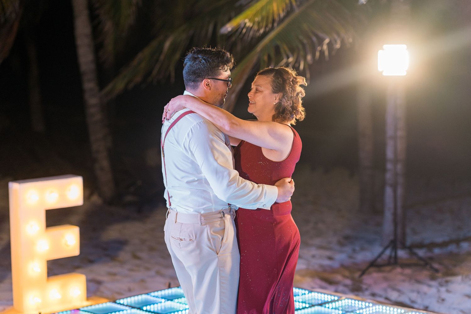 Couple dancing elegantly on LED dance floor beside illuminated letter E at beach wedding.