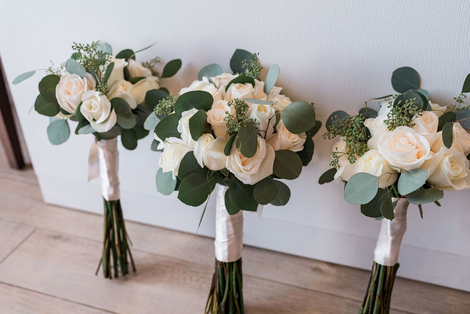 Three elegant bridal bouquets with white roses and eucalyptus greenery tied with white ribbon.