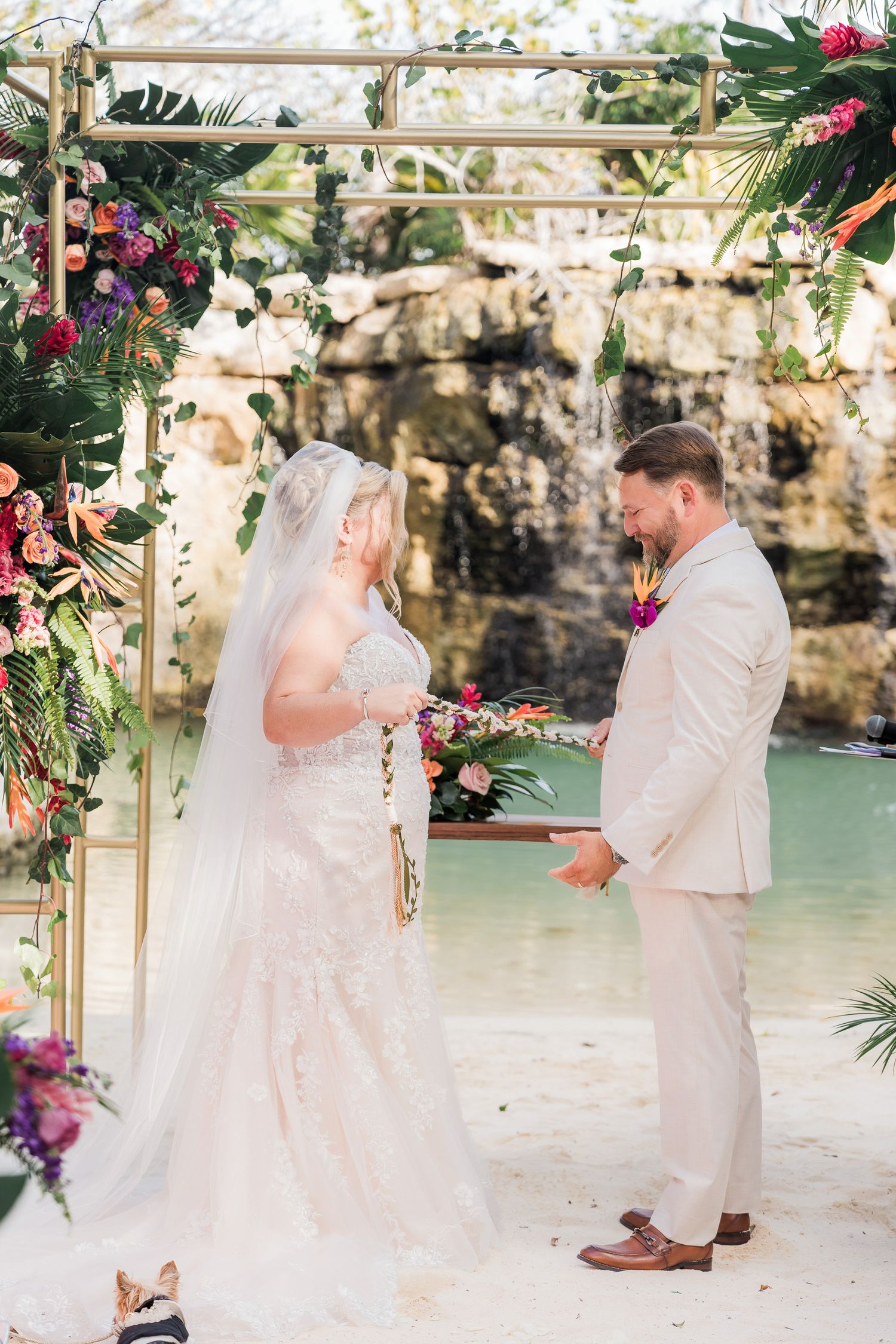 Hotel Xcaret México Wedding  - Bride and groom exchange vows under a floral arch by a waterfall at a tropical beach wedding ceremony.