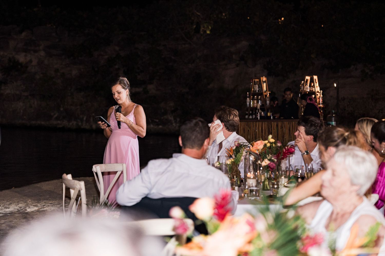 Woman in pink dress giving a speech at an elegant outdoor wedding reception dinner at night.