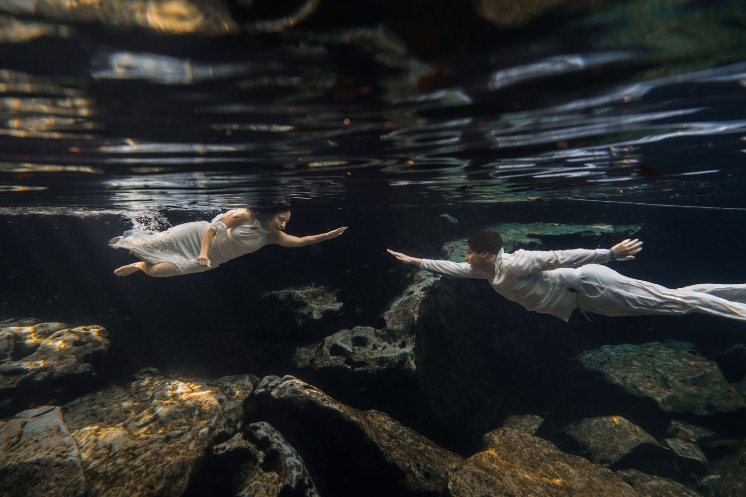 Two people in white dresses swim gracefully underwater among rocks and fish in clear dark water.