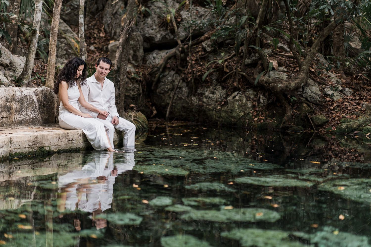 Couple in white outfits sitting by a serene jungle cenote with lily pads and lush greenery in Mexico.