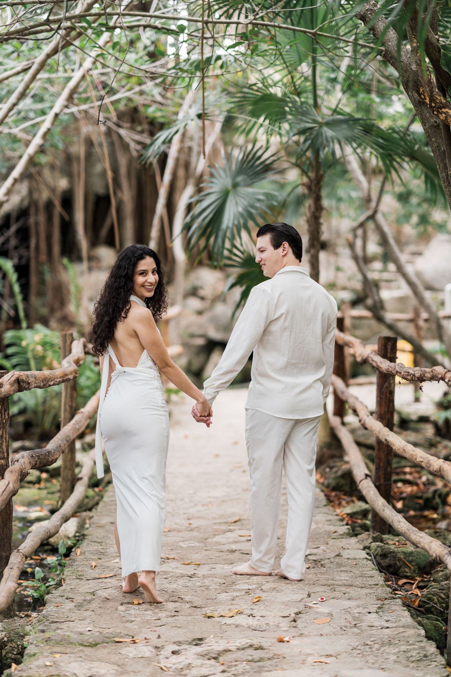 Couple in white attire holding hands on a rustic wooden bridge surrounded by lush tropical greenery.