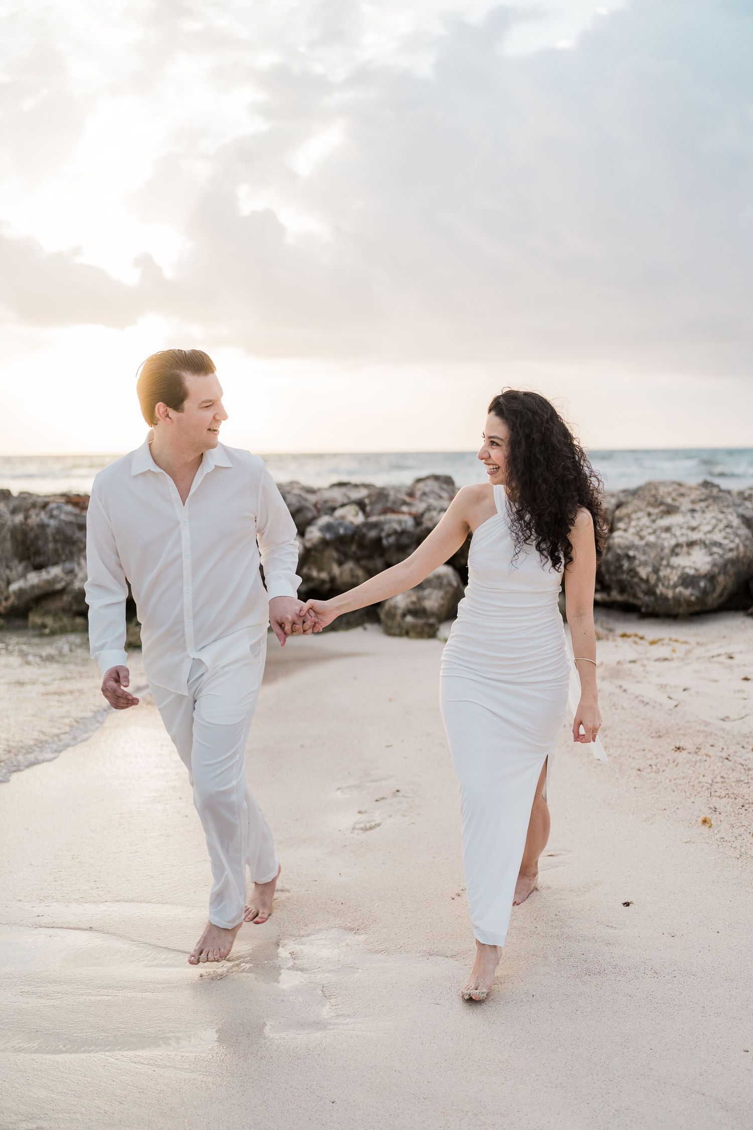 Couple in white outfits holding hands and walking barefoot on a sandy beach at sunset with rocky shore behind them.