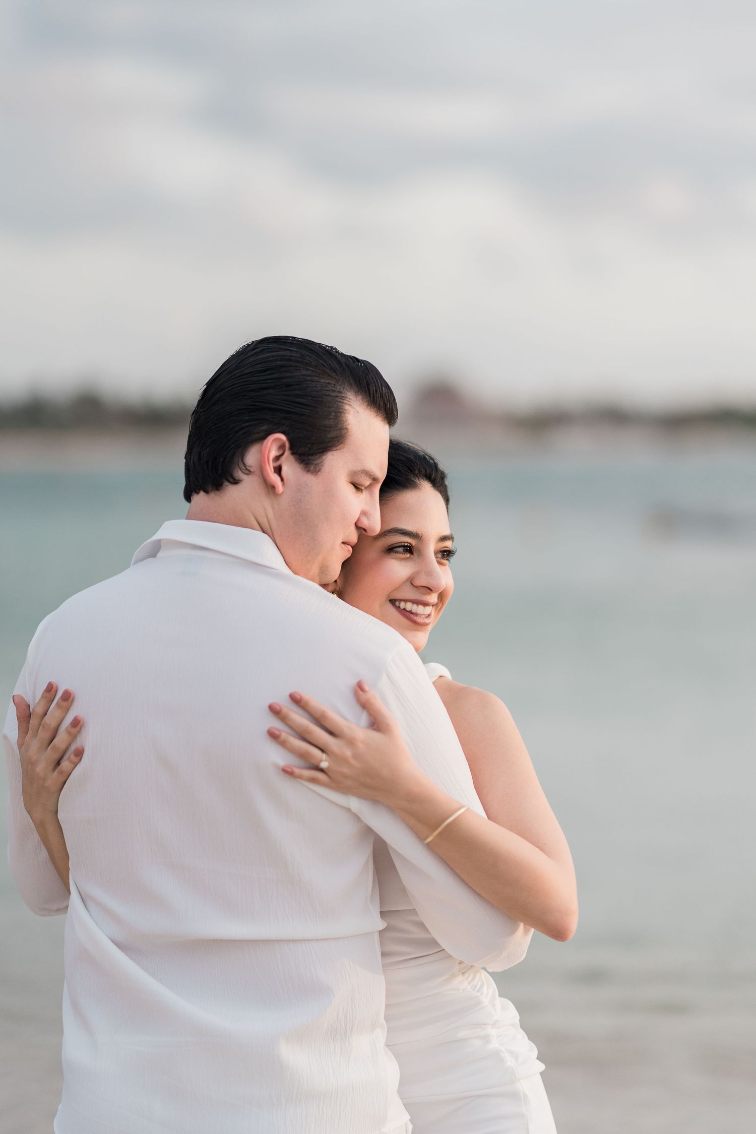 A smiling bride in white embraces her groom on a beach, with calm turquoise water in the background.