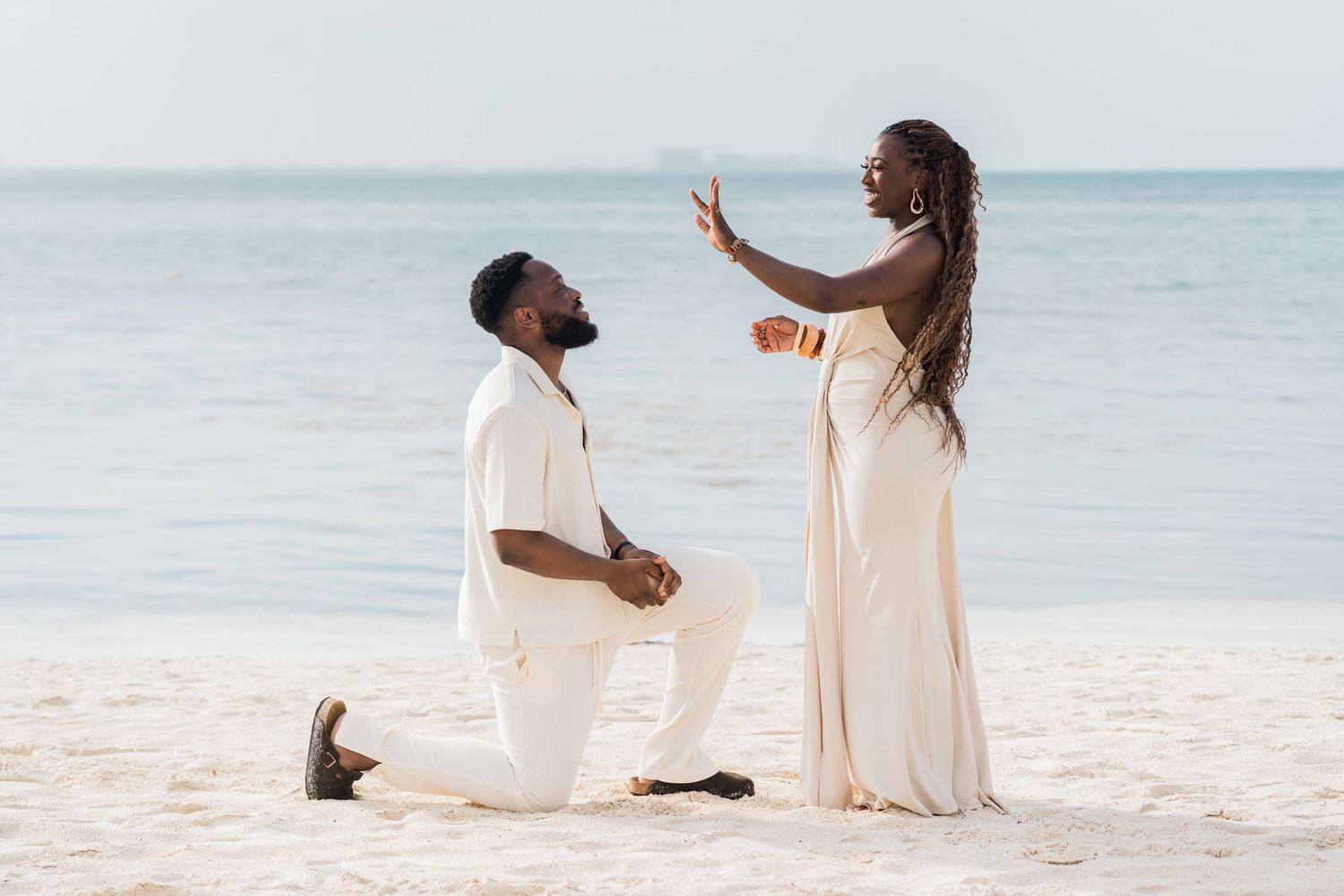 Beach marriage proposal with man kneeling as woman raises hand in surprised reaction by the ocean.