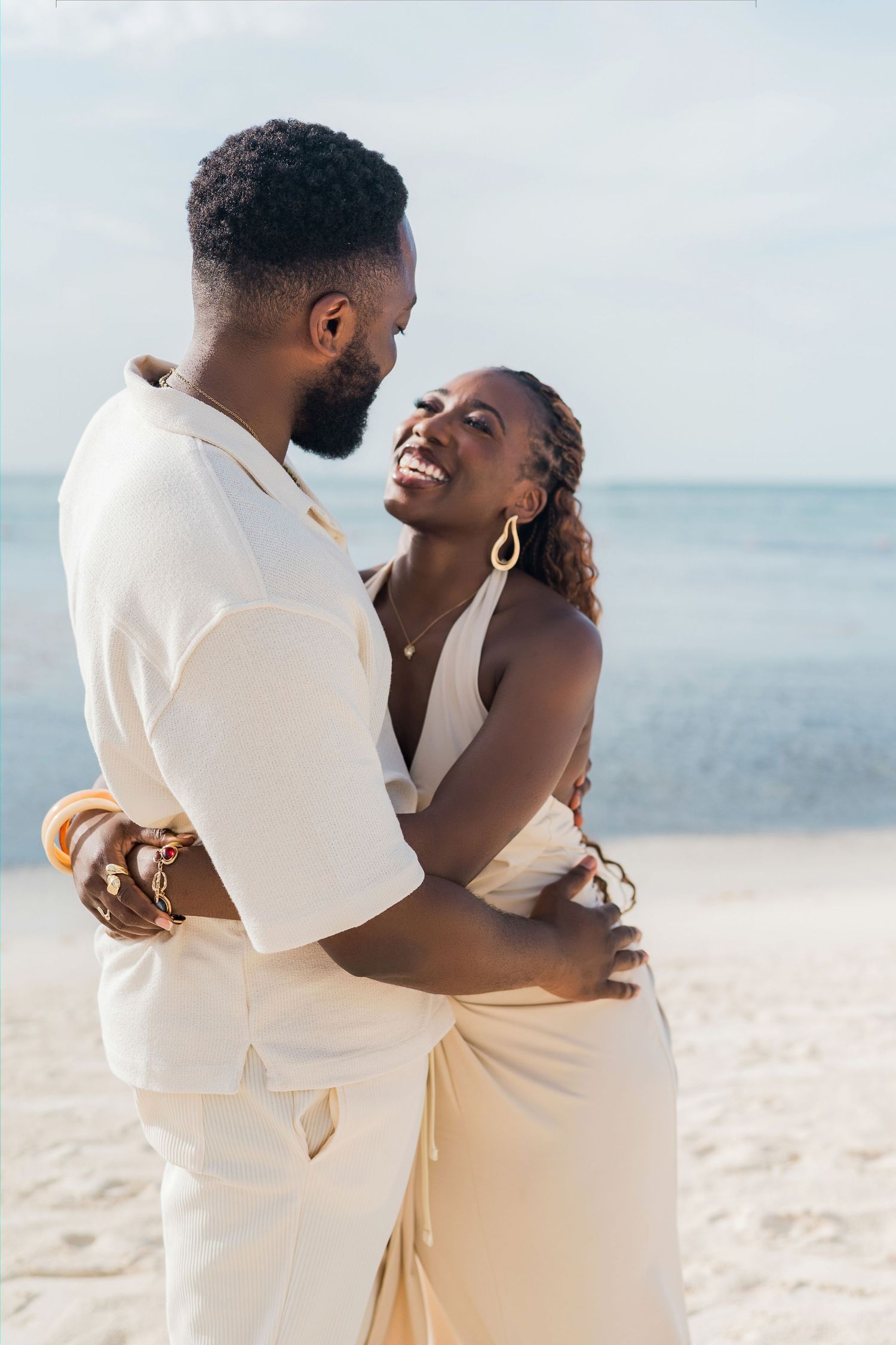 A happy couple embraces on a sandy beach, both dressed in elegant white and cream outfits, smiling at each other.