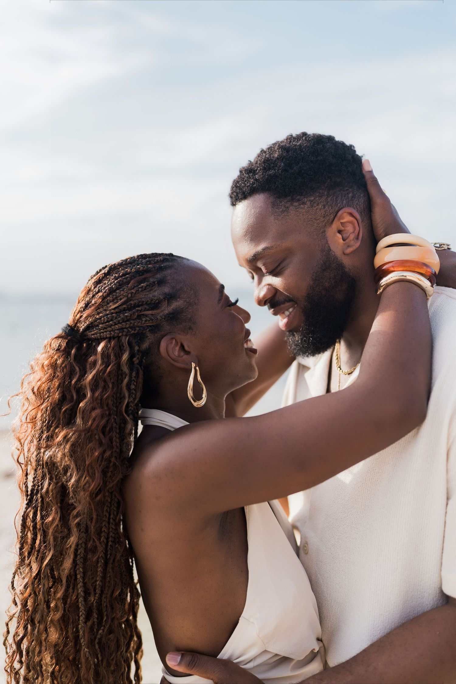 A romantic Black couple embraces on a beach, the woman with braids holding the man's neck as they lean in close.