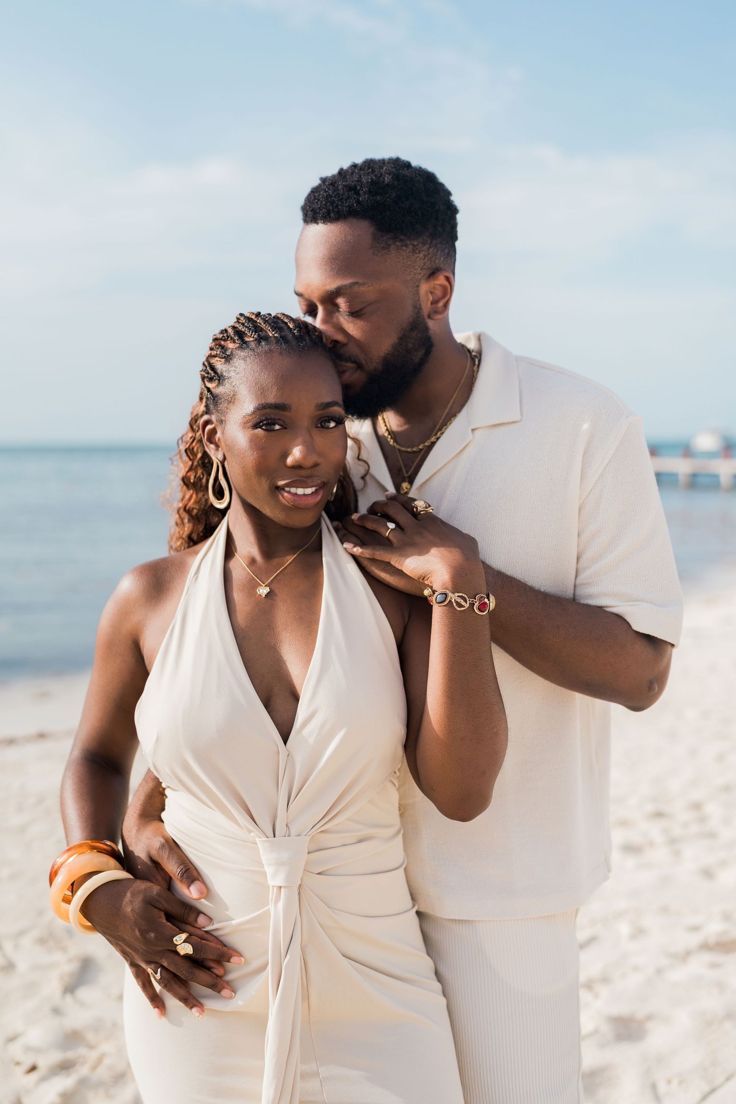 A smiling Black woman in a white halter dress stands with her partner on a sunny beach, showcasing gold jewelry and braided hair.