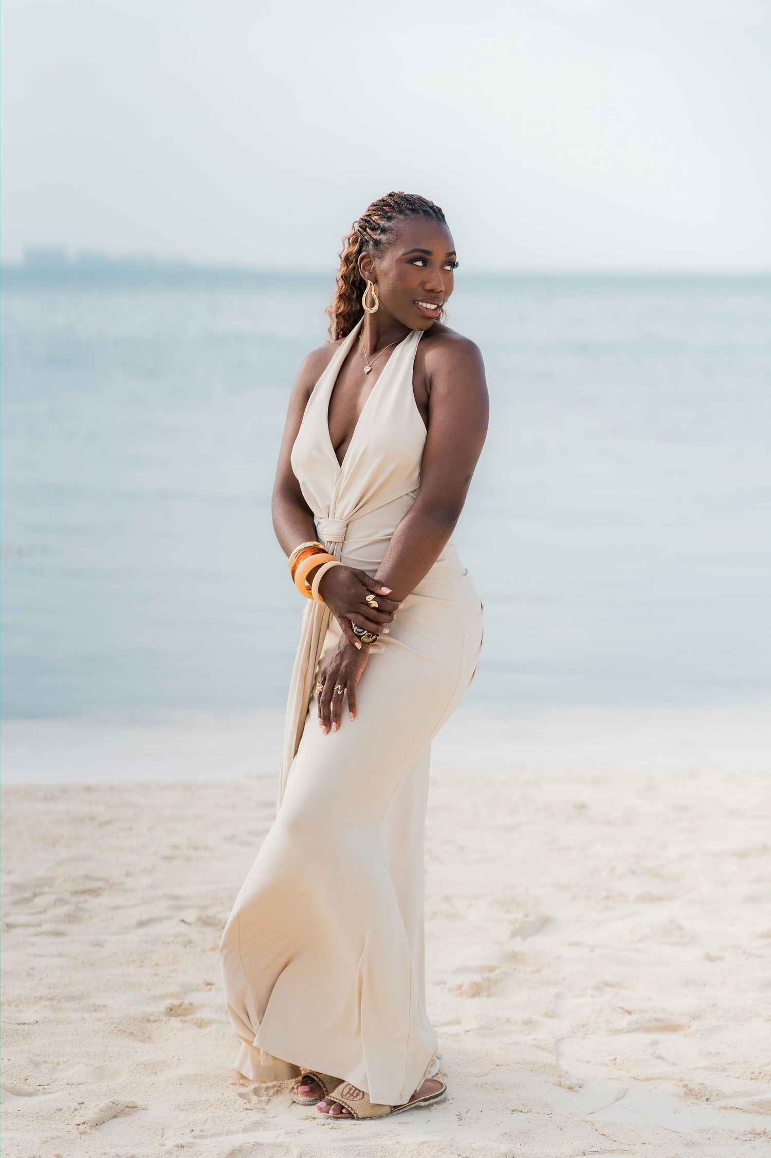Elegant Black woman in cream halter maxi dress poses on sandy beach with ocean backdrop, wearing gold jewelry.