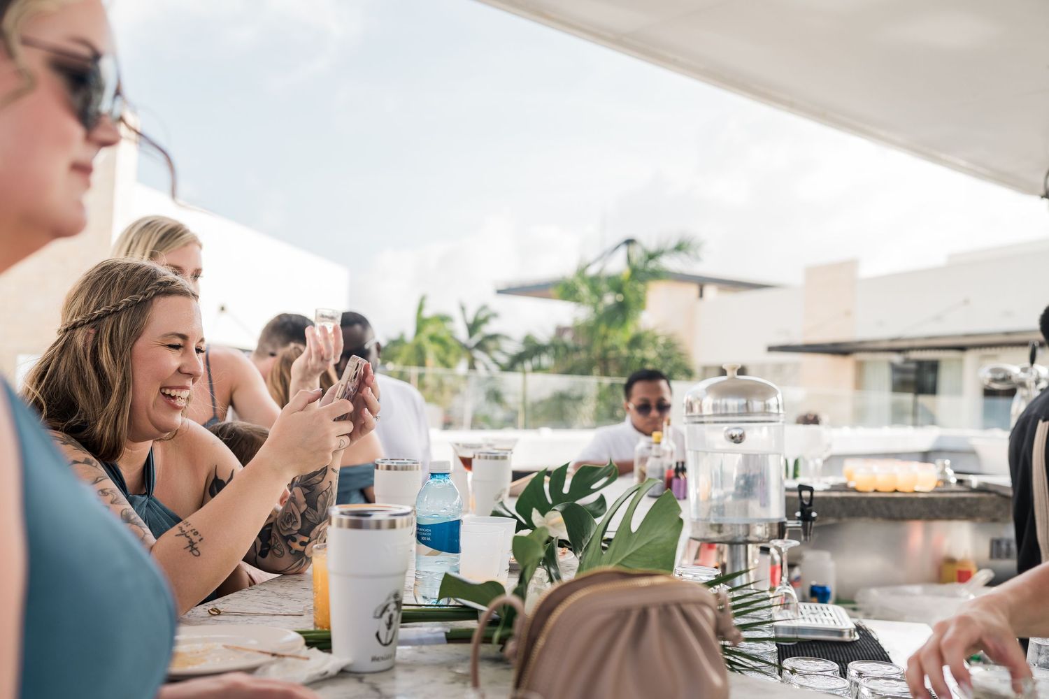 Women laughing and taking photos at an outdoor rooftop party with tropical decor and beverages.