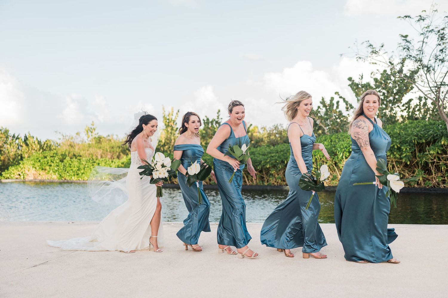 Bride and bridesmaids in teal gowns walk on a sandy beach with lush greenery and water in the background.