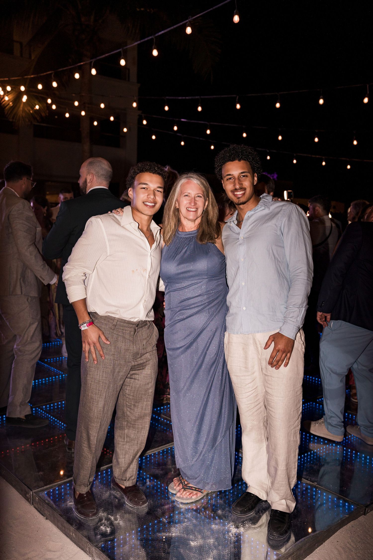 Three people smiling together at an outdoor evening event with string lights, standing on a reflective dance floor.