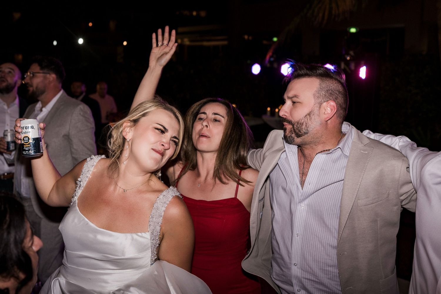 Three wedding guests dancing joyfully at a nighttime outdoor reception, woman in white bridal gown raises hand.