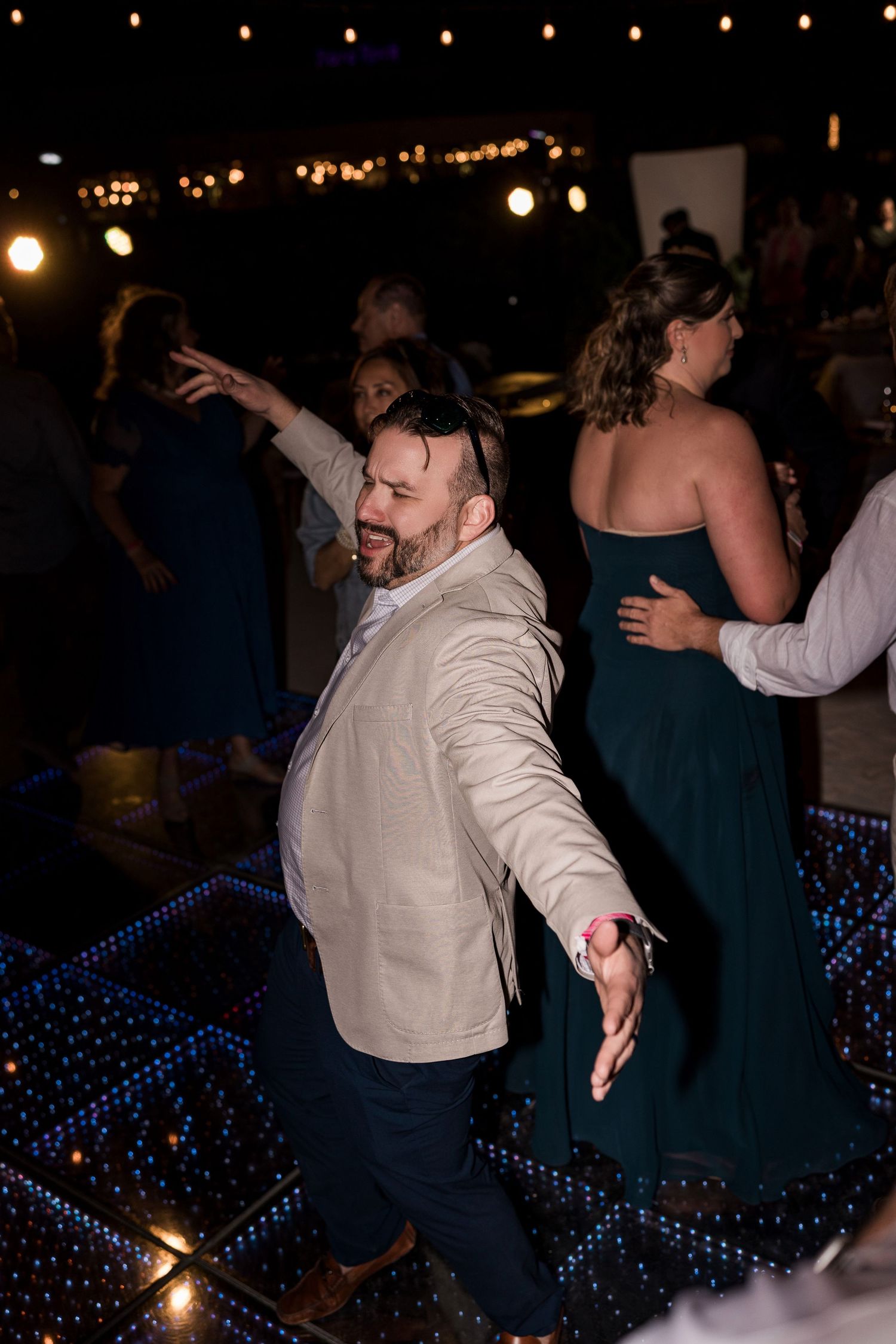 Man in beige blazer dancing energetically on a lit dance floor at a wedding reception at night.