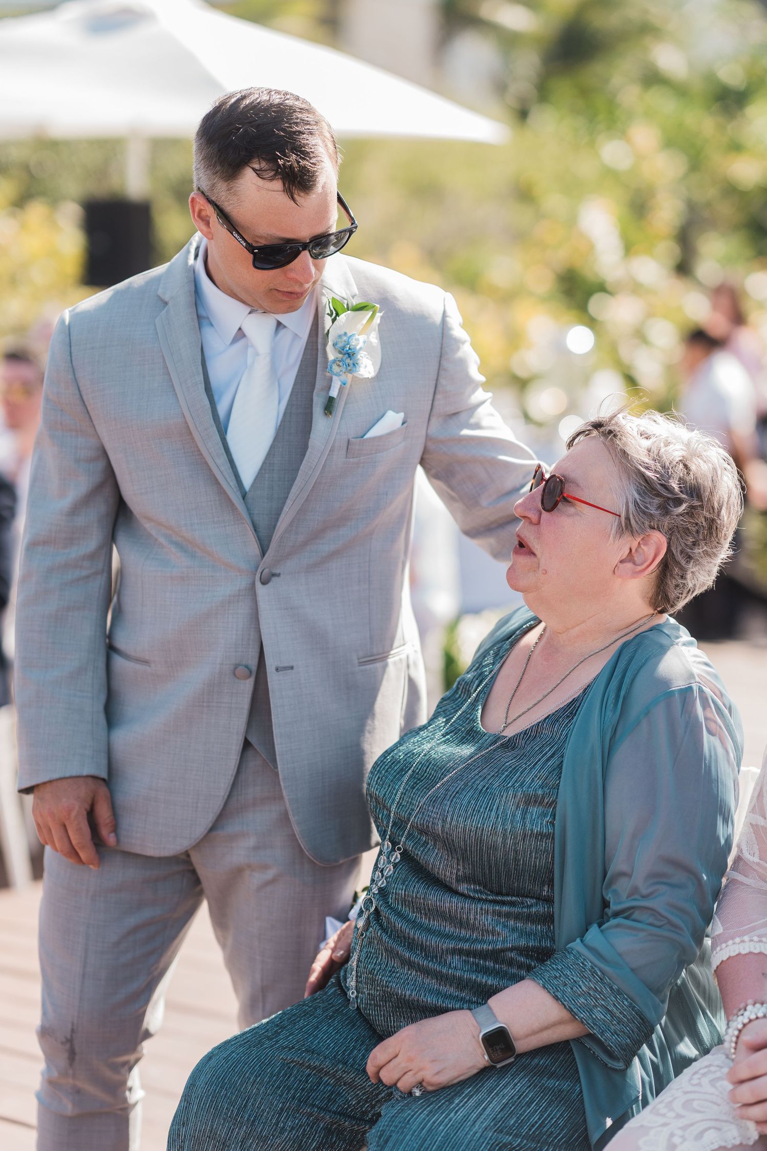 Groom in gray suit with boutonniere speaks to seated older woman in teal dress at outdoor wedding ceremony.