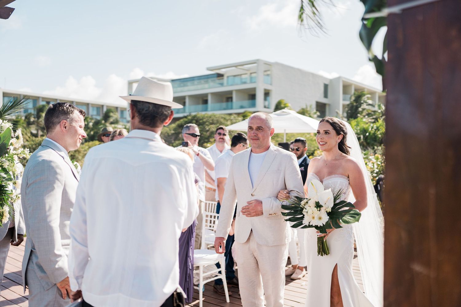 Bride in white gown walks with escort at outdoor beach wedding ceremony with guests seated nearby.