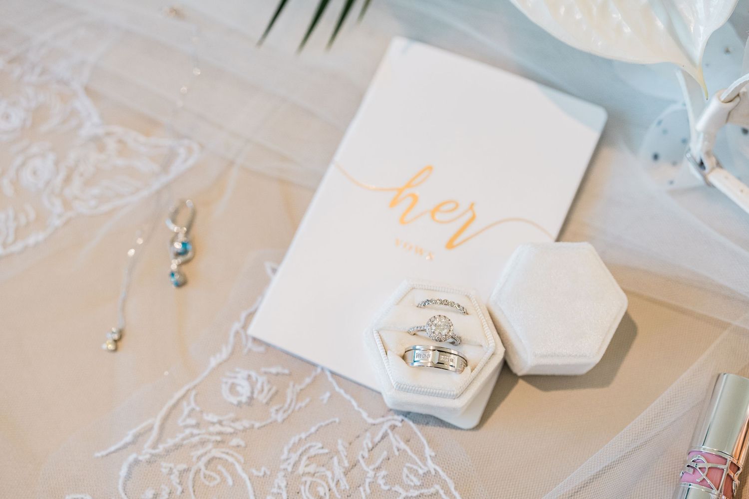 Elegant wedding flat lay with rings in marble hexagon box, her vow book, and delicate jewelry on lace fabric.