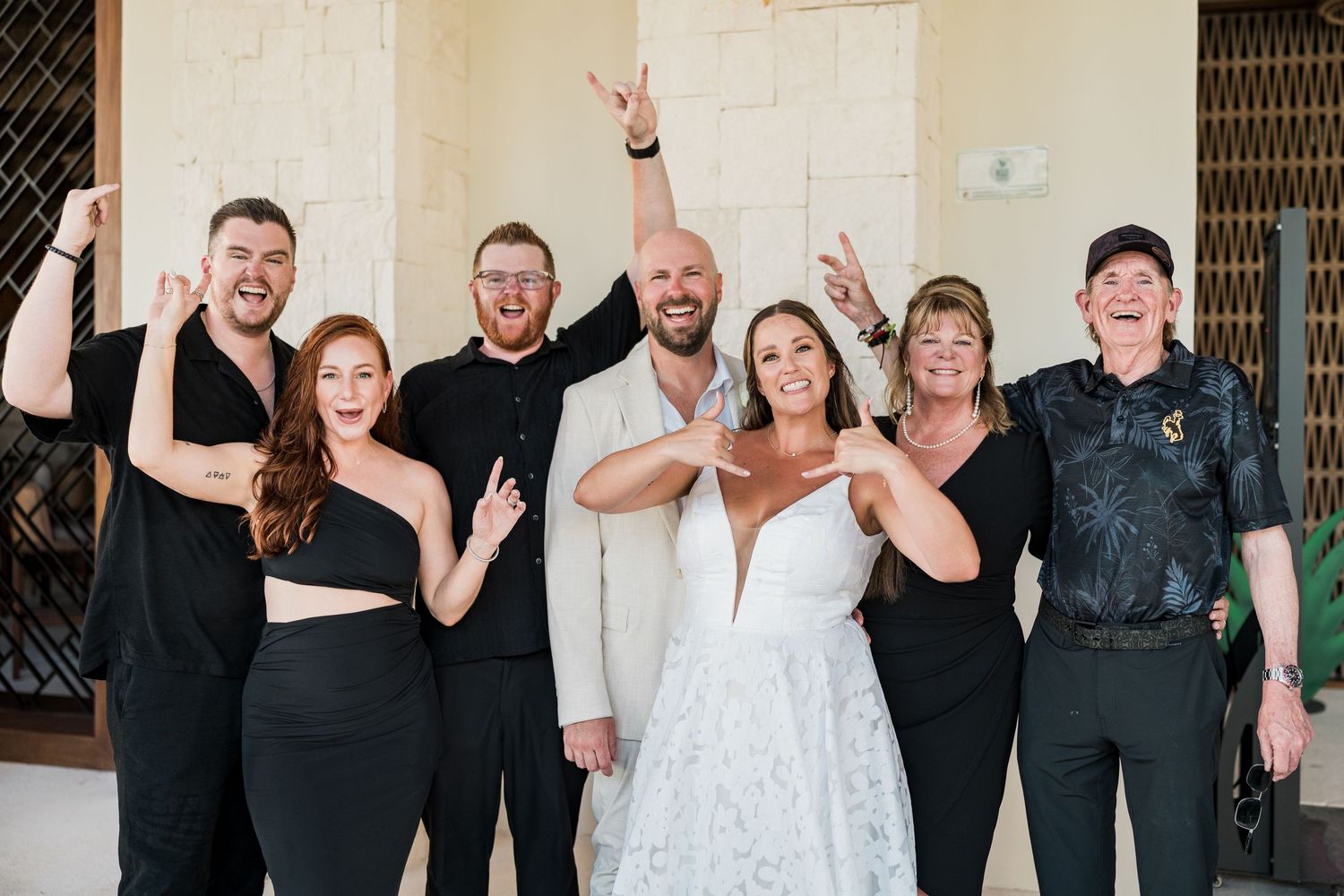 Wedding party in black attire celebrates with bride and groom in white, posing joyfully outdoors.