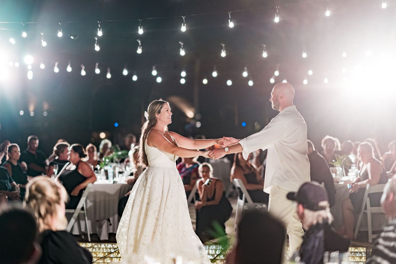 Bride and groom share first dance under string lights at outdoor evening wedding reception with guests watching.