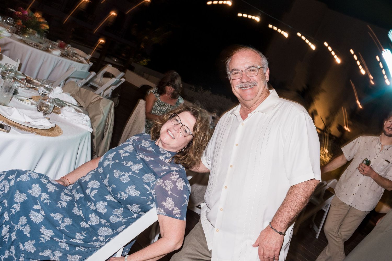 Smiling couple posing together at an outdoor wedding reception with festive string lights in background.