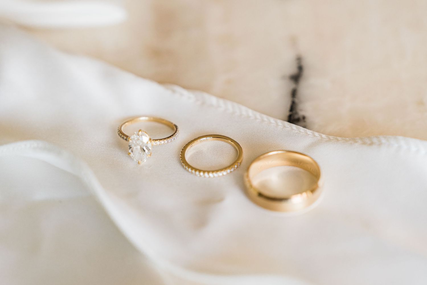 Three gold wedding rings on white fabric, including a diamond engagement ring, eternity band, and plain wedding band.