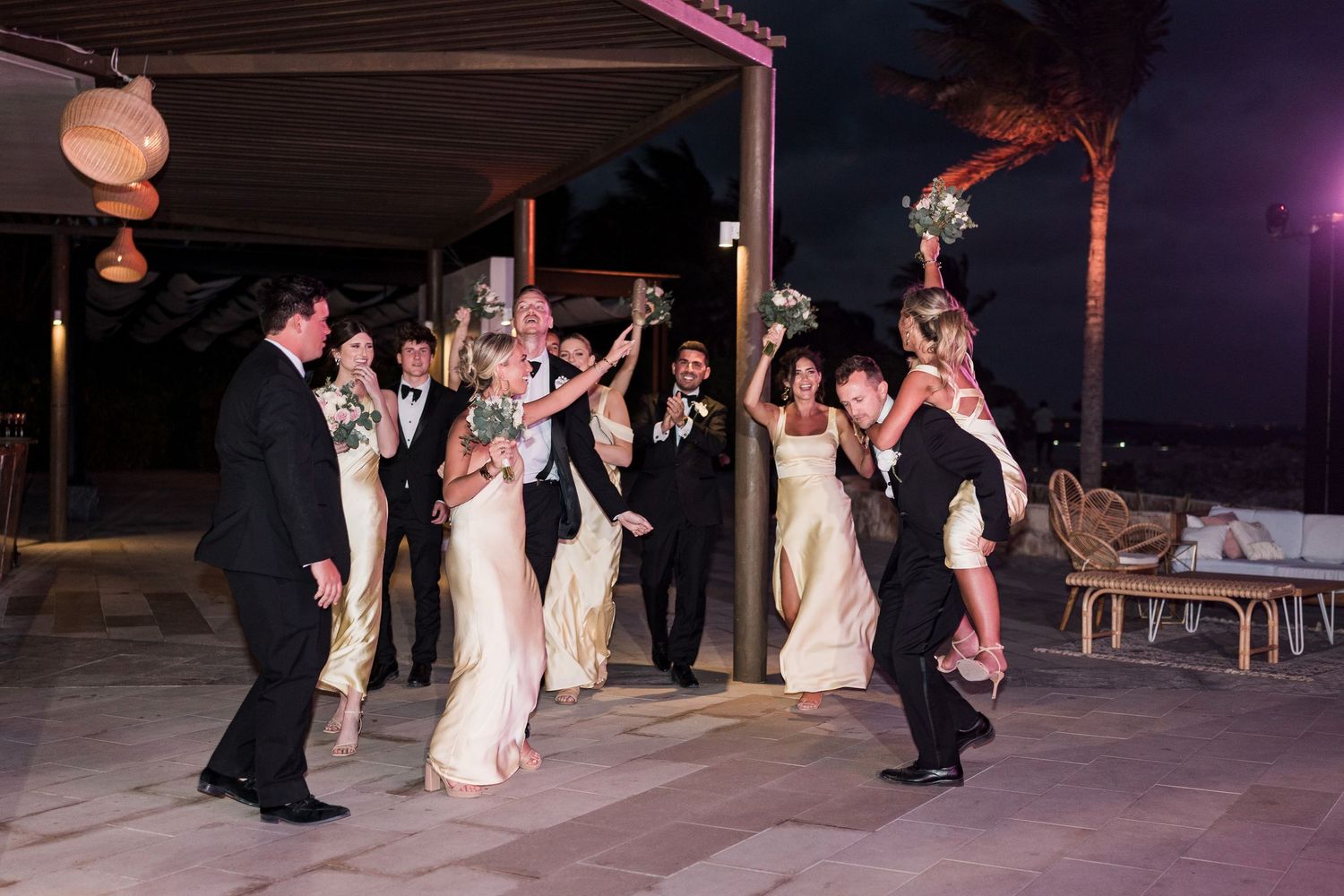 Wedding party celebrating outdoors at night, bridesmaids in champagne gowns and groomsmen in tuxedos cheering with bouquets raised.