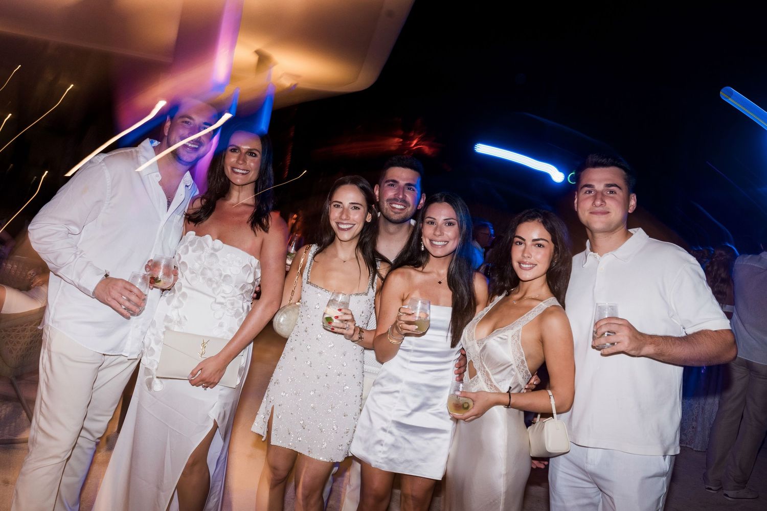 Group of friends in white outfits celebrating at an outdoor party, holding drinks and smiling.
