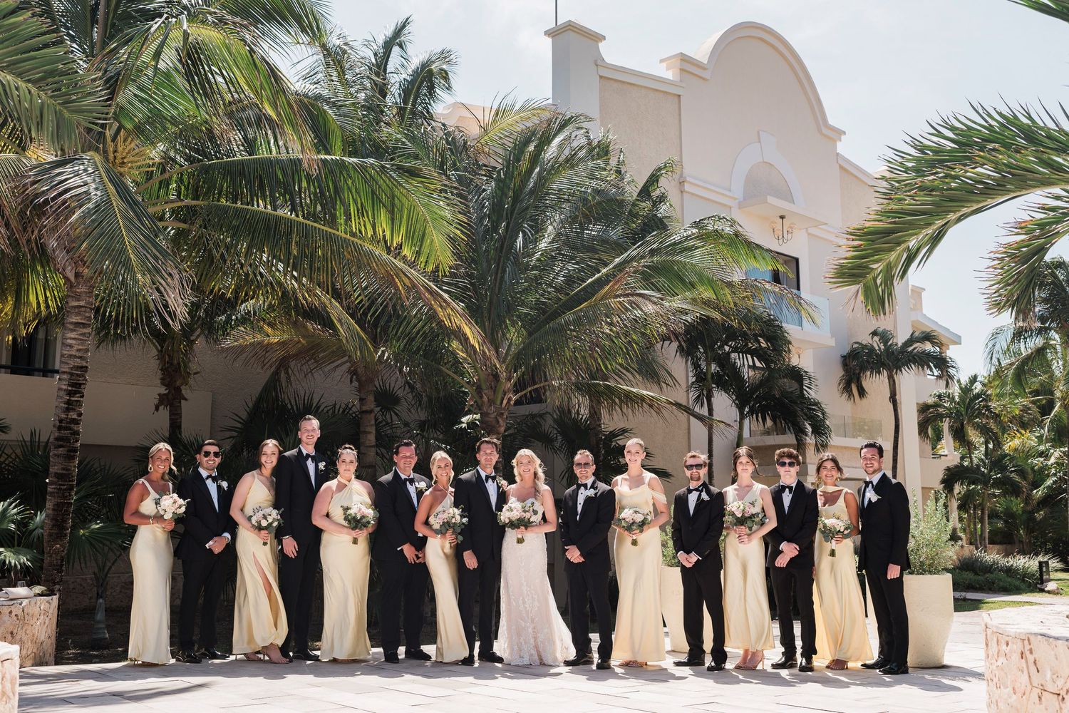 Wedding party in formal attire poses outdoors with palm trees and white building backdrop at tropical resort venue.