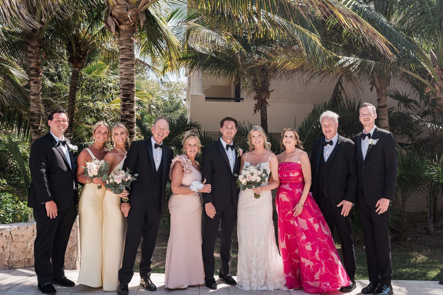 Wedding party poses outdoors near palm trees, wearing formal attire including tuxedos and elegant gowns.