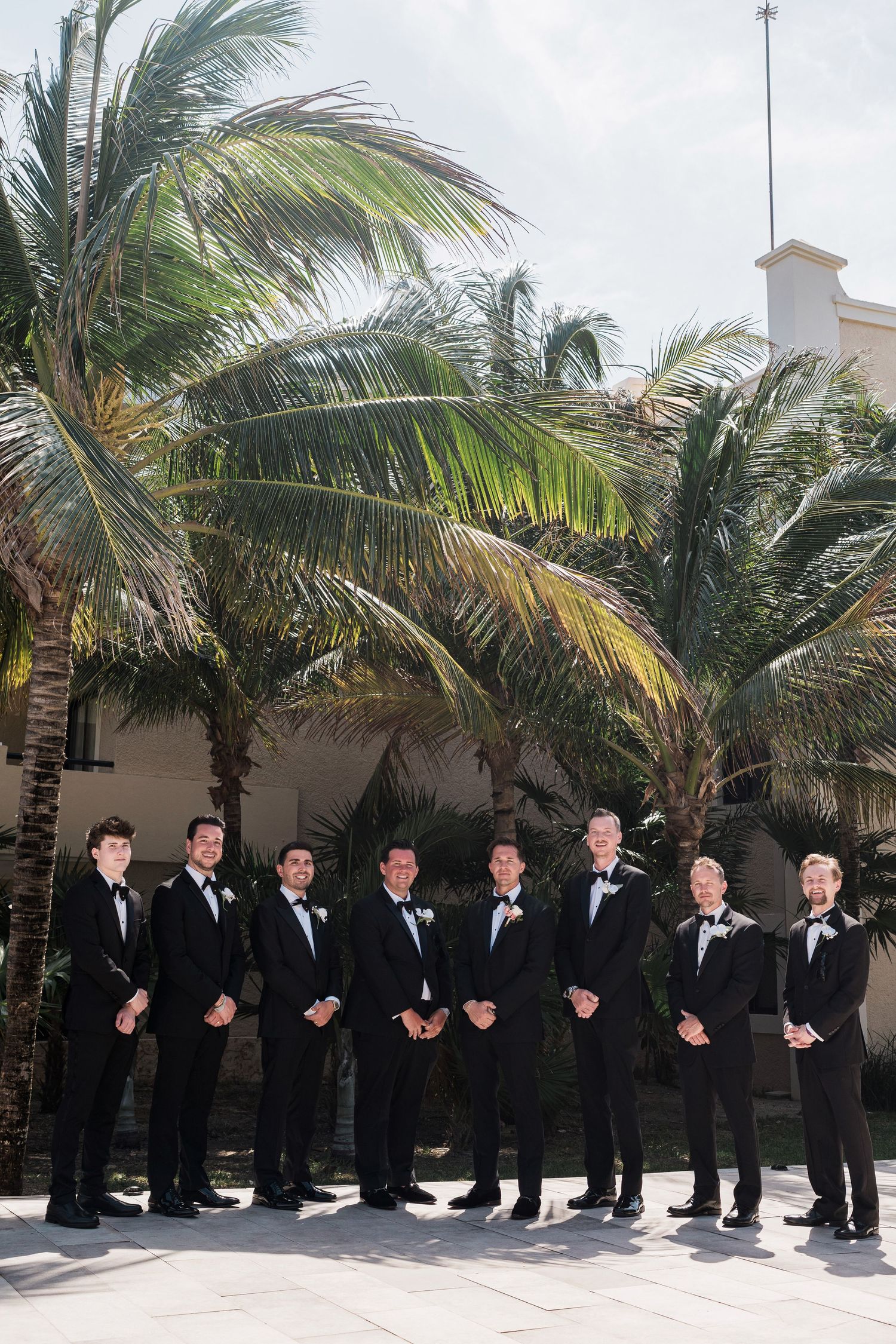 Groomsmen in black tuxedos with bow ties pose together outdoors near tropical palm trees at a destination wedding.