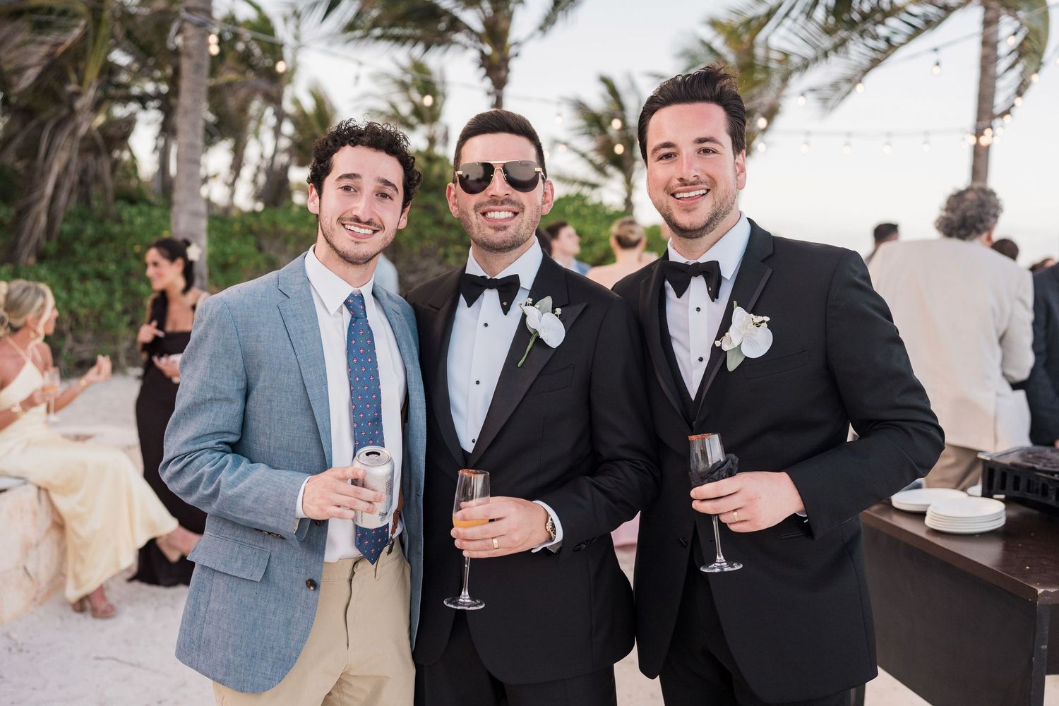 Three men in formal attire smiling and holding drinks at a beachside wedding reception.