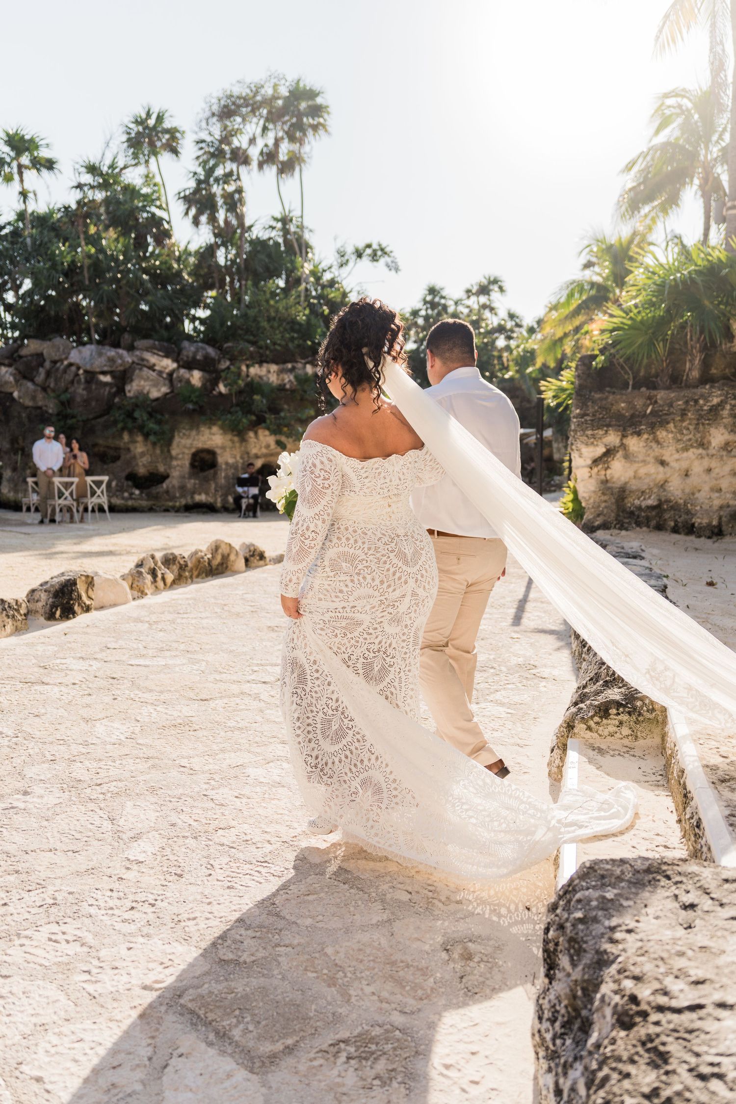 Xcaret Arte Wedding - Bride in lace gown with flowing veil walks with groom in tan suit at outdoor beach wedding ceremony.