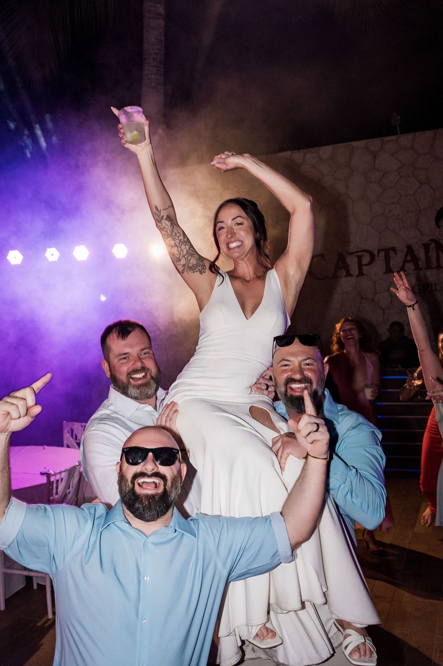 Joyful bride in white dress lifted by groomsmen on festive dance floor with purple lights.