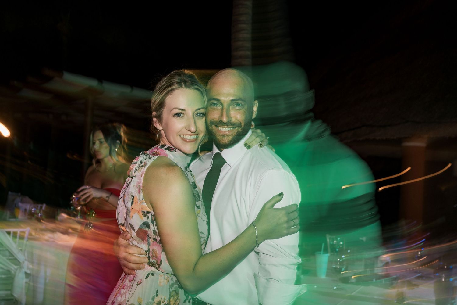 A smiling couple embraces while dancing at an outdoor evening wedding reception with green lighting.