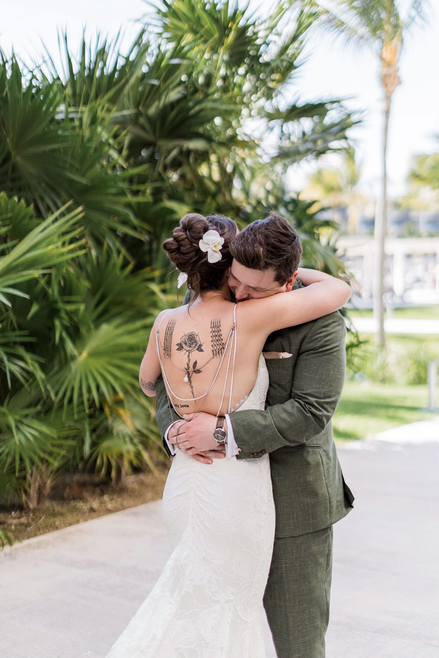 Bride and groom embrace in front of lush tropical palms, bride wearing backless lace gown with floral tattoos.