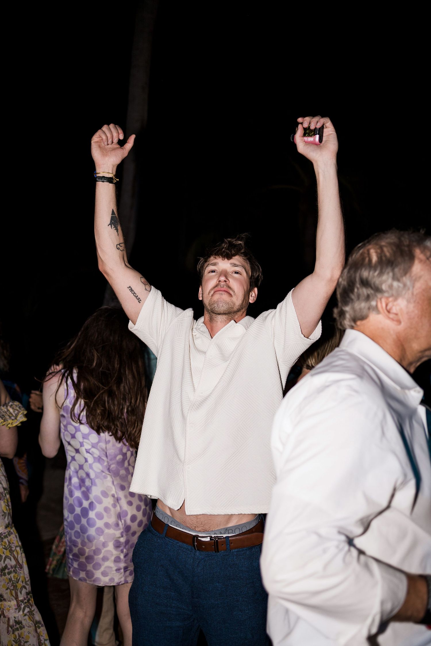Man with tattoos dancing with arms raised at a nighttime outdoor party event.