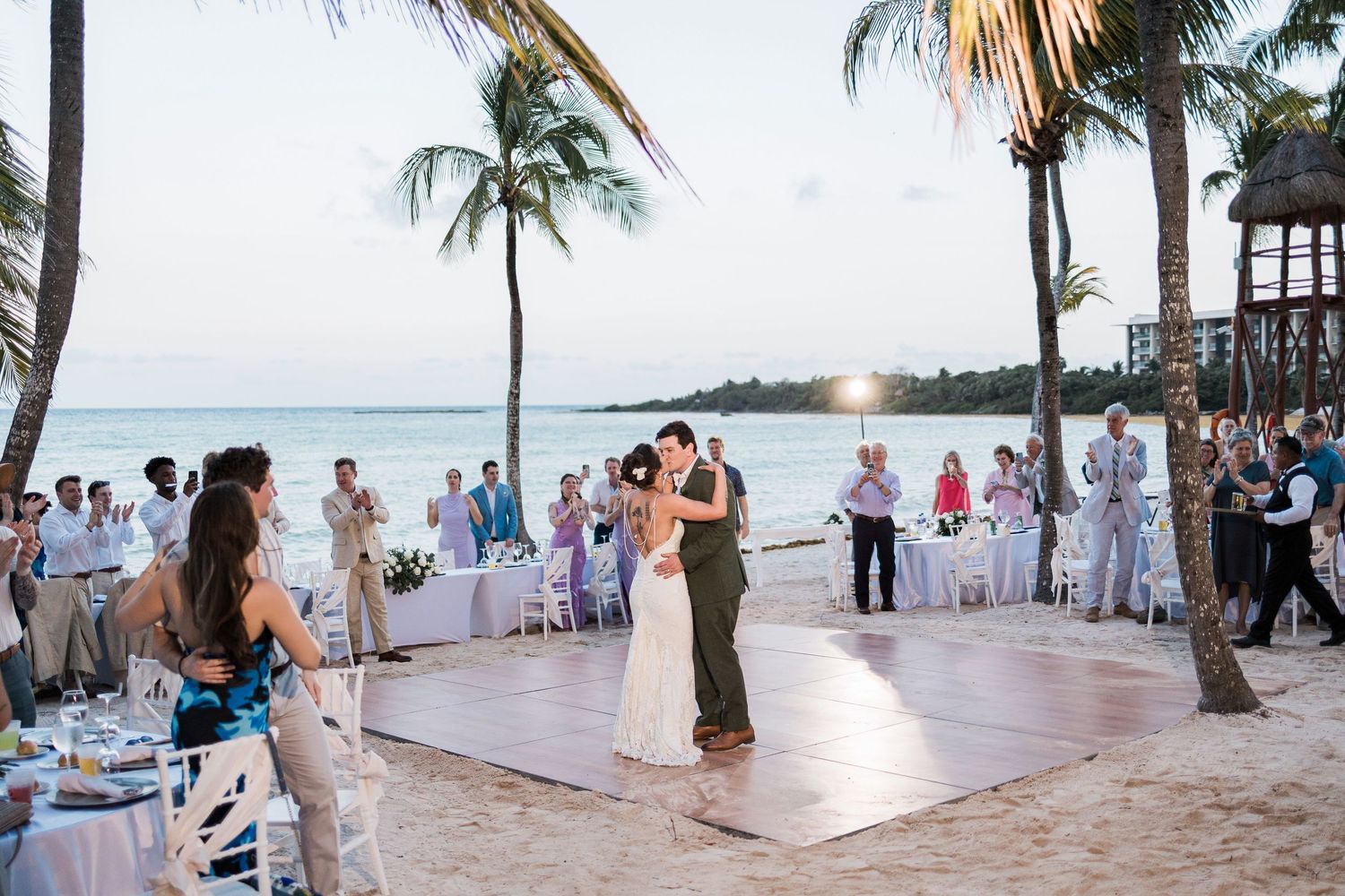 Couple shares first dance on beach wedding reception dance floor at sunset with palm trees and guests.