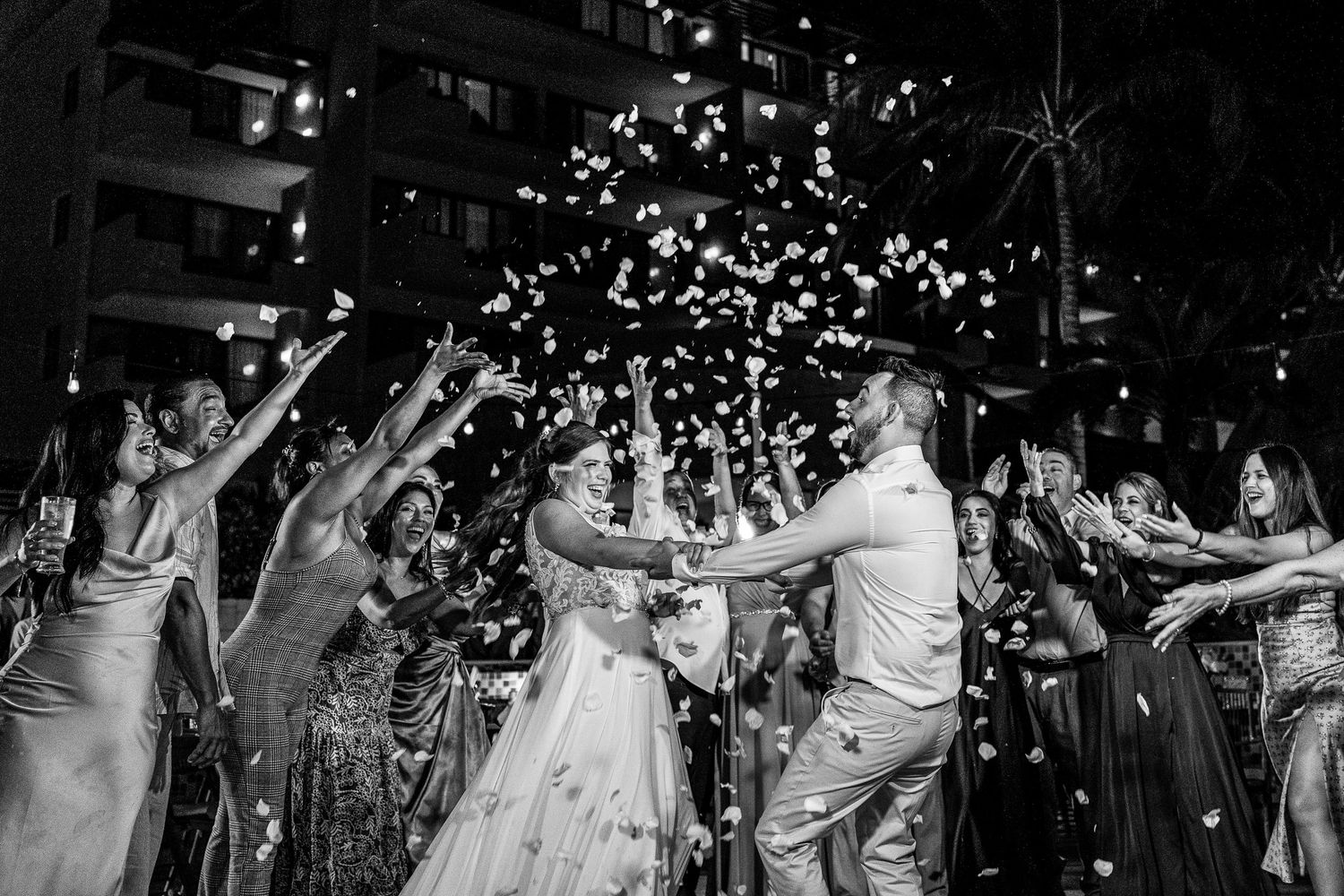 Bride and groom celebrate joyfully as guests toss confetti during festive wedding reception in black and white photo.