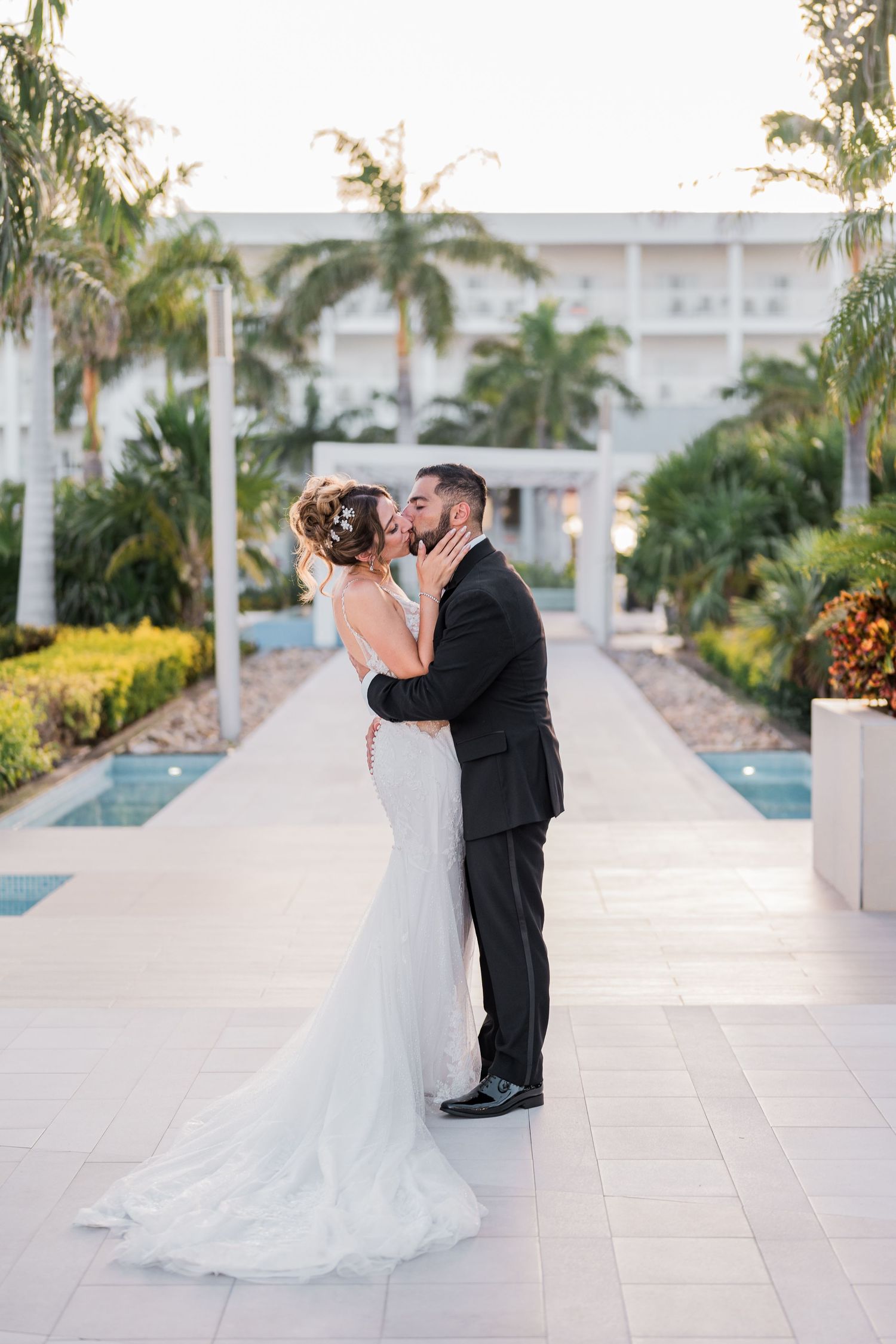 Bride and groom share a romantic kiss at a tropical resort with palm trees.